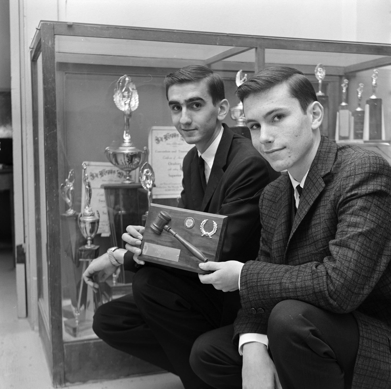 Black and white photo of debaters crouching in front of a trophy case holding their trophy