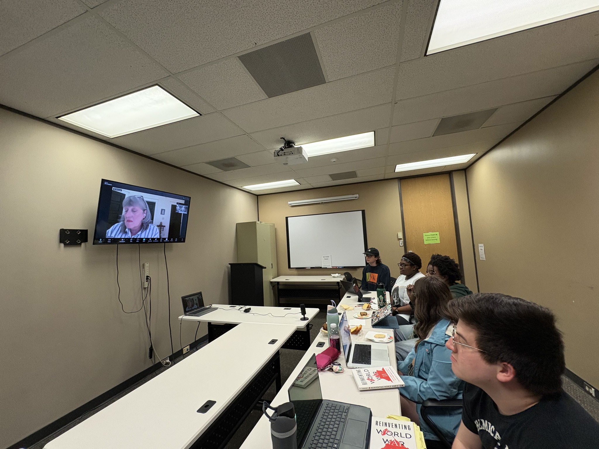 Students sitting at table look up to TV featuring Barbara Biesecker on Zoom meeting