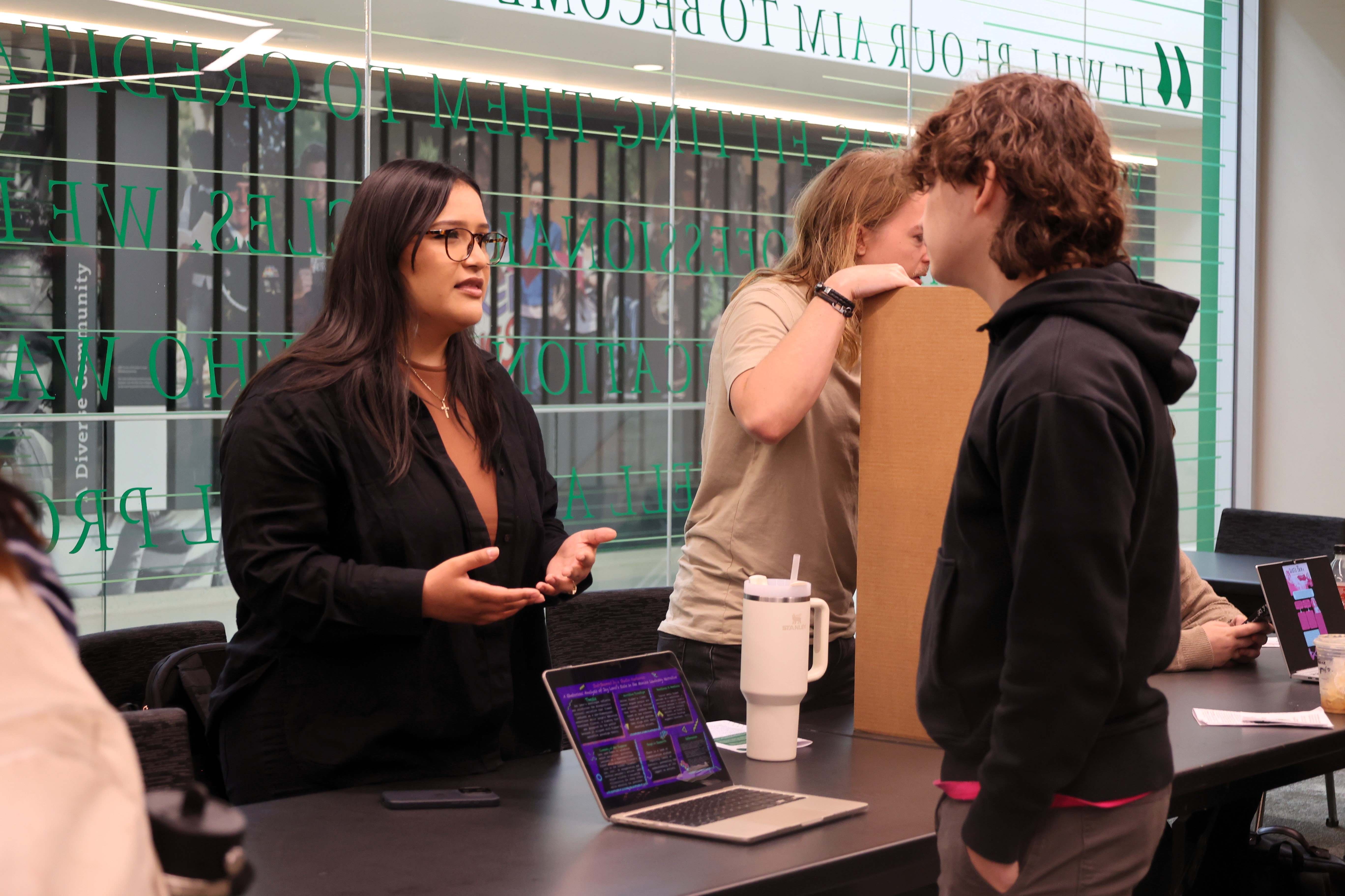 Student standing behind table with laptop giving presentation to a student standing in front of the table