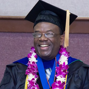 UNT COMM Alum Samuel Muwanguzi in black cap and gown with a lei of purple and white flowers on his neck